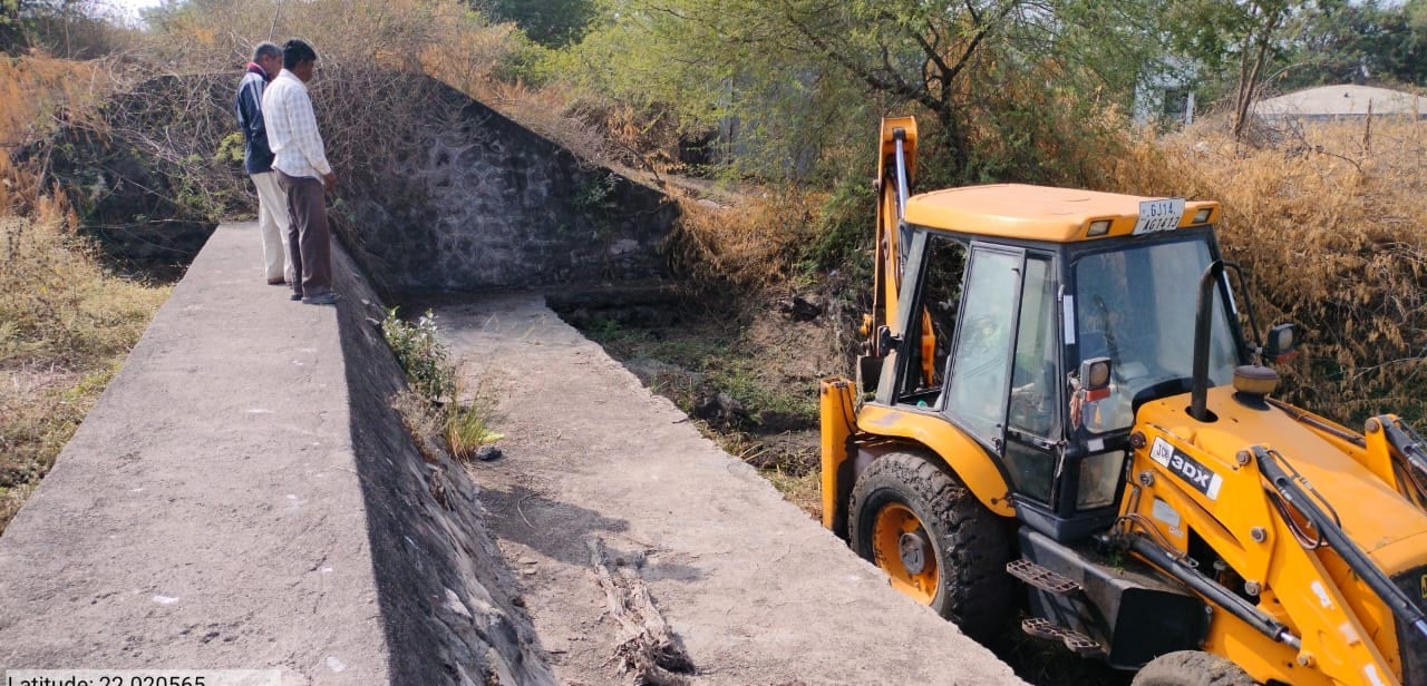A JCB excavator deepening the silted bed behind a concrete check dam while two men watch from the wall above