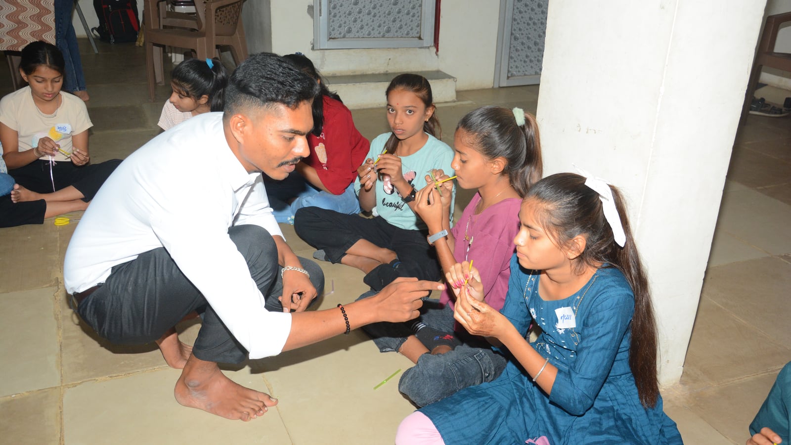 A facilitator crouching among children seated on the floor, showing them how to fold a drinking straw into a working reed whistle