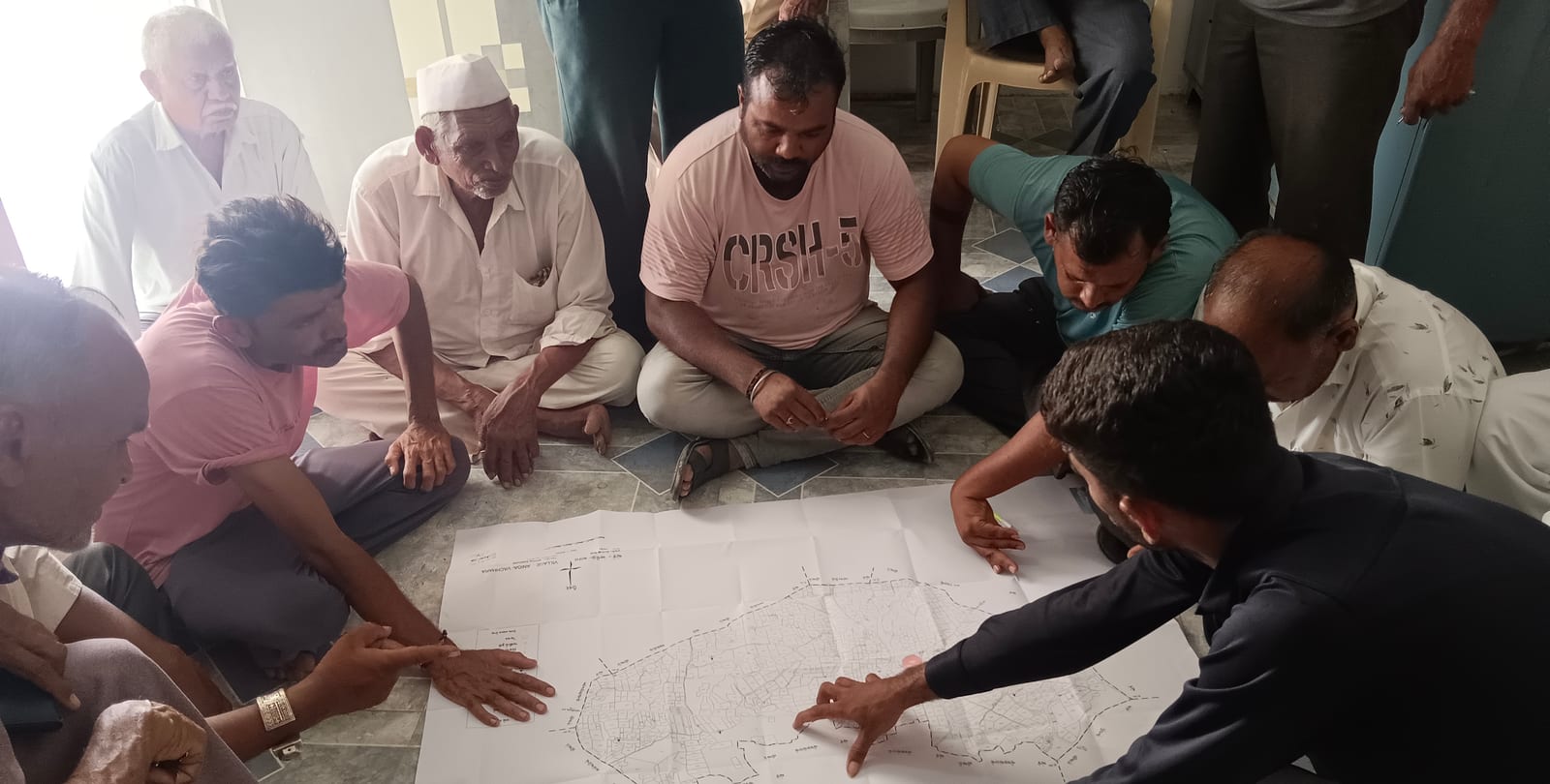 Men from the village seated on the floor around a large cadastral map, tracing survey boundaries with their hands