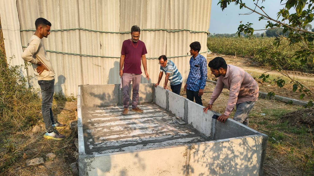 A newly built concrete vermicompost tank beside a sheet-metal shed, with five men inspecting the finished walls
