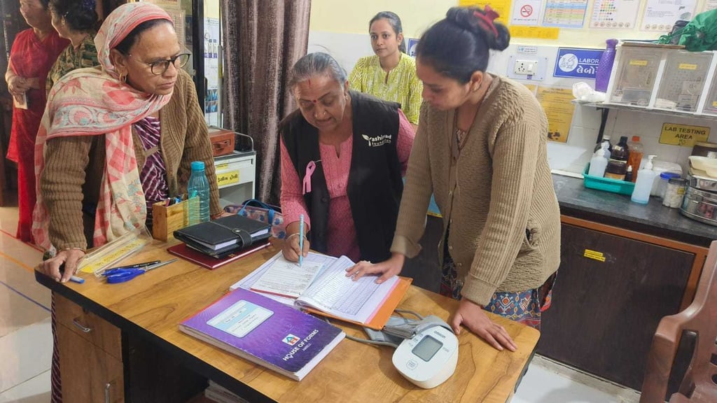 A health worker filling in a screening register at a camp desk, a blood pressure monitor beside her, two women waiting at the desk