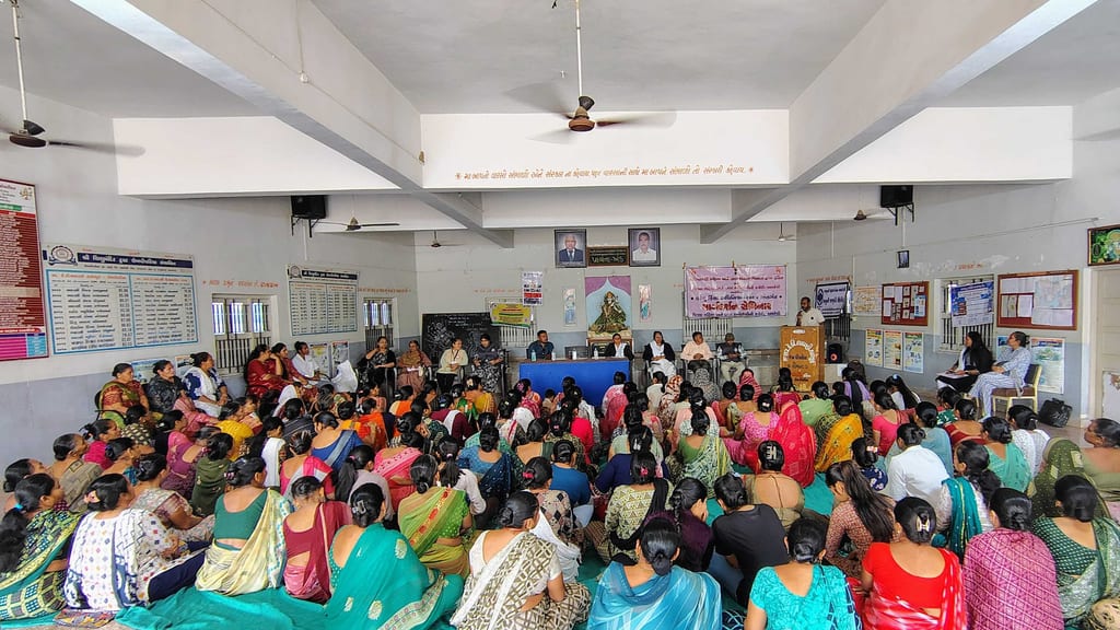 A hall full of women seated on the floor at a guidance seminar, a speaker at the microphone under a banner naming the Domestic Violence Act