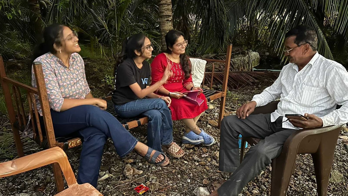 Three students sitting on wooden chairs in a farmyard talking with a farmer, one of them taking notes